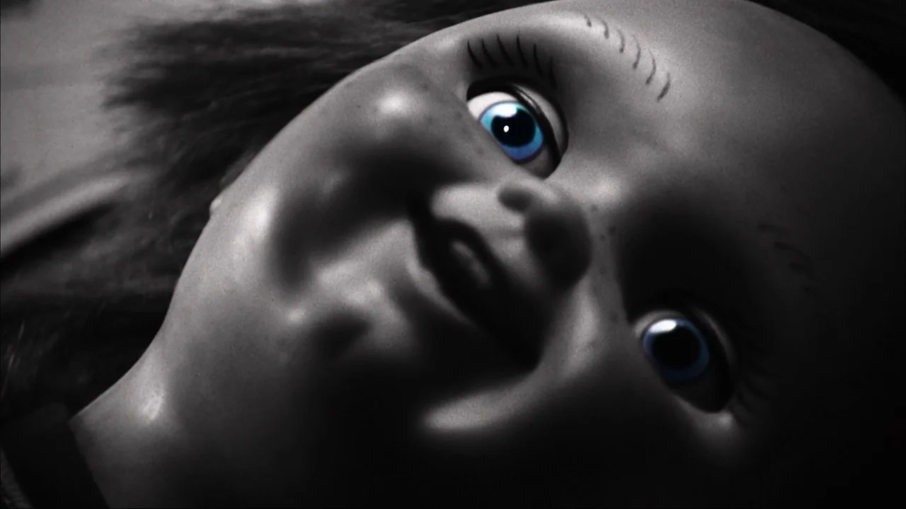 Curse of Chucky (2013) movie still: closeup — A doll face occupies the frame in a close-up against a dark backdrop. The doll maintains a neutral,…
