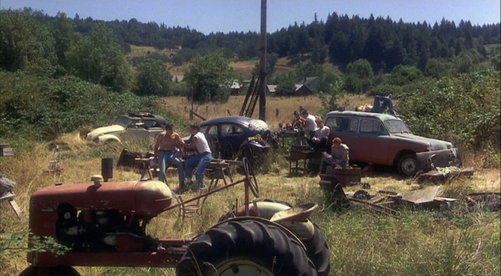 Stand By Me (1986) movie still: wide — An outdoor lot contains several rusted vintage cars and a large red farm tractor. Young adults stand and sit…