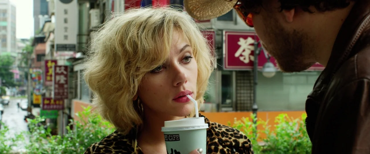 Lucy (2014) movie still: medium — A street in Taipei features blurred buildings with red signs and lush green plants. A blonde woman holding a paper…