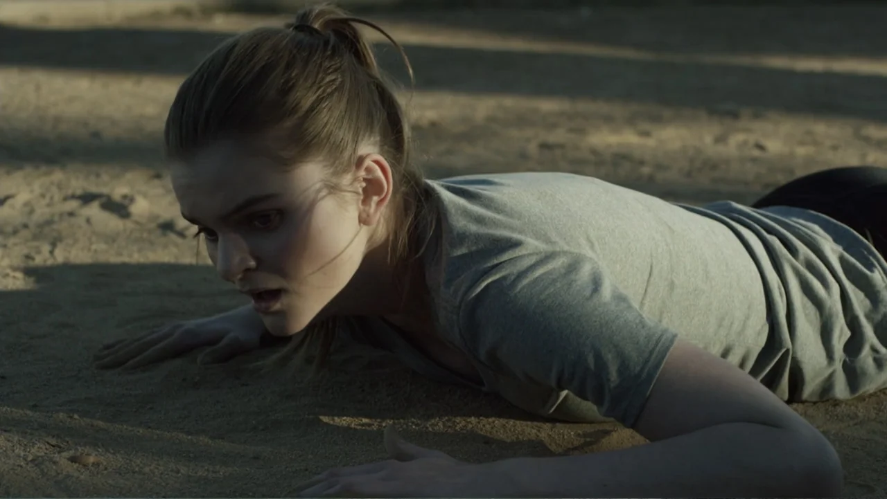 Totem (2022) movie still: medium — An open expanse of sand creates a flat, textured ground surface. A woman lies prone on the sand, resting on her…