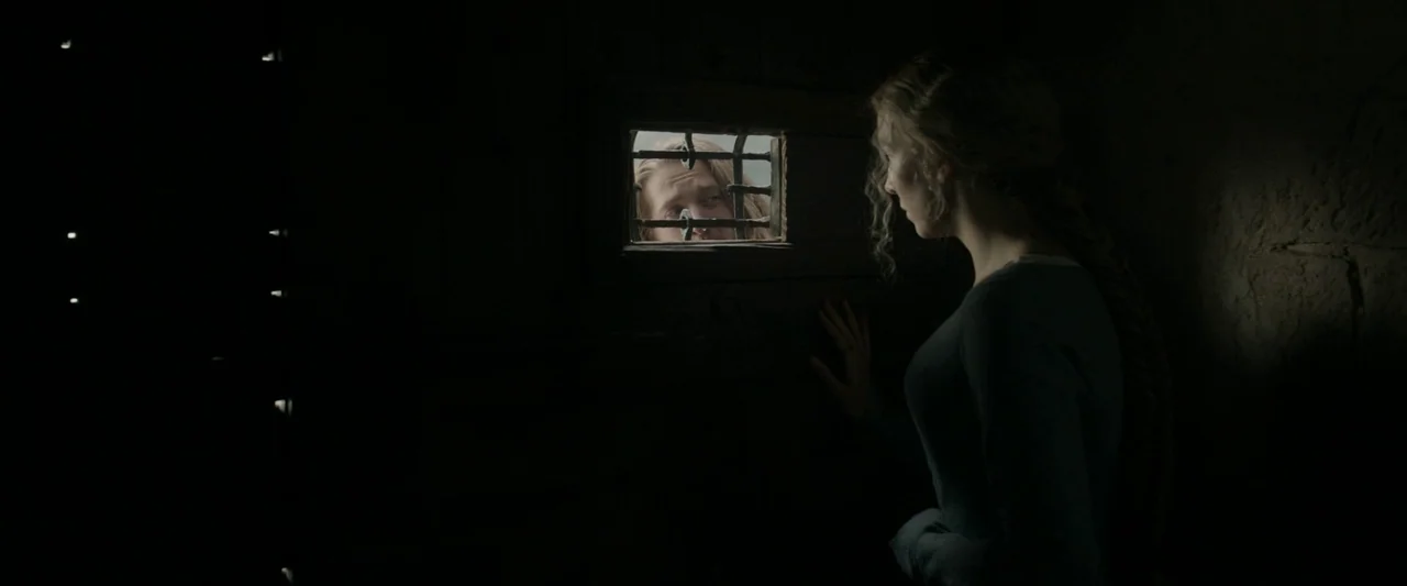 The Last Duel (2021) movie still: medium — A dimly lit room with heavy timber walls and a small barred viewing portal. A woman faces the portal,…