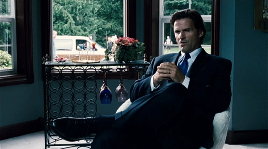 Visioneers (2008) movie still: medium — A well-lit room features large windows showing a green, wooded exterior and a car. A man in a suit sits in a…