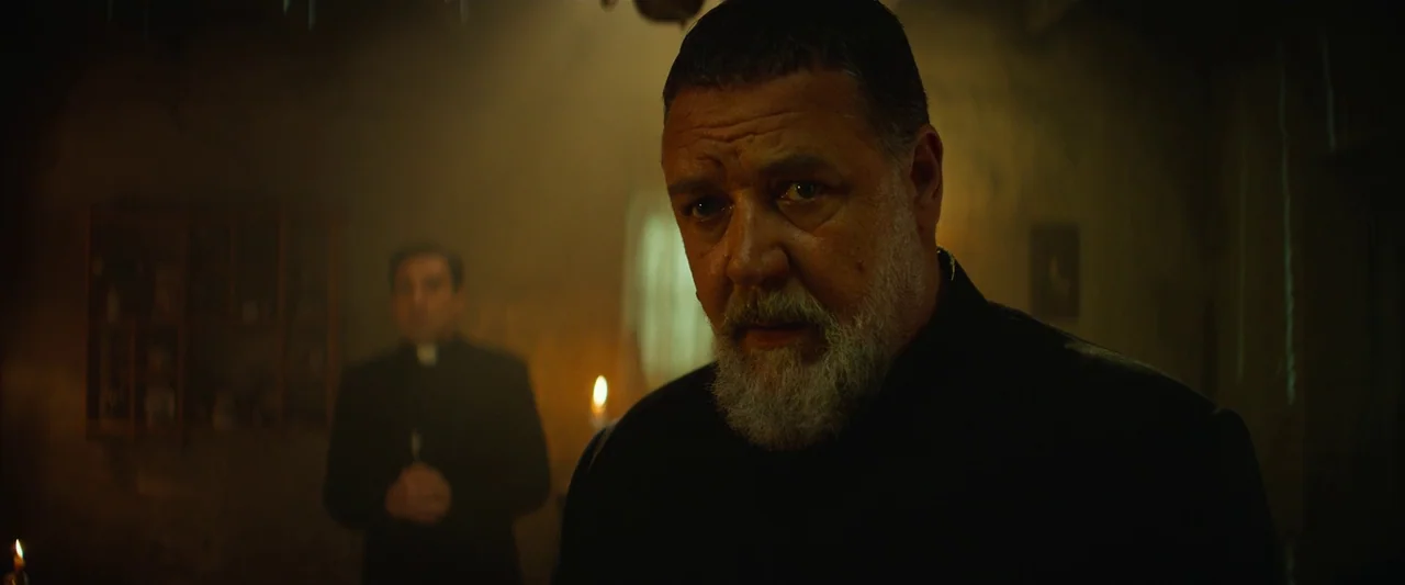 The Pope’s Exorcist (2023) movie still: closeup — A dimly lit room with stone walls and scattered candles providing minimal light. A bearded priest…