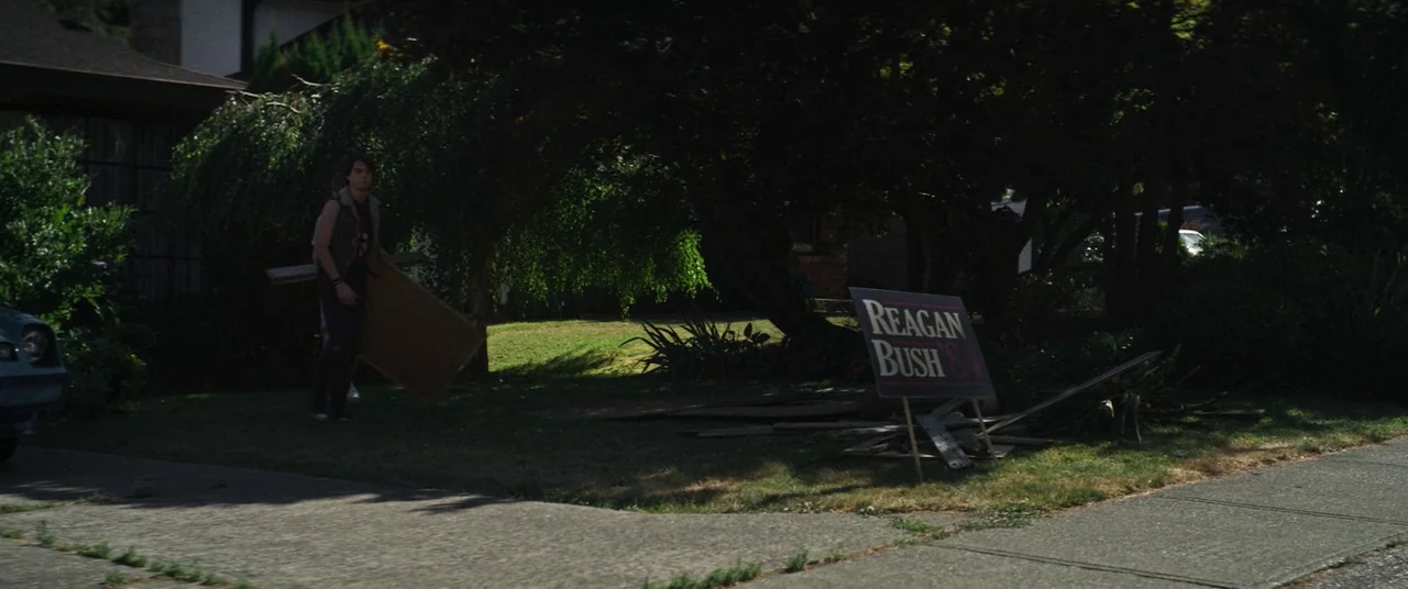 Summer of 84 (2018) movie still: wide — A suburban front yard features a large tree and a political yard sign. A teenage boy carries a wooden board…