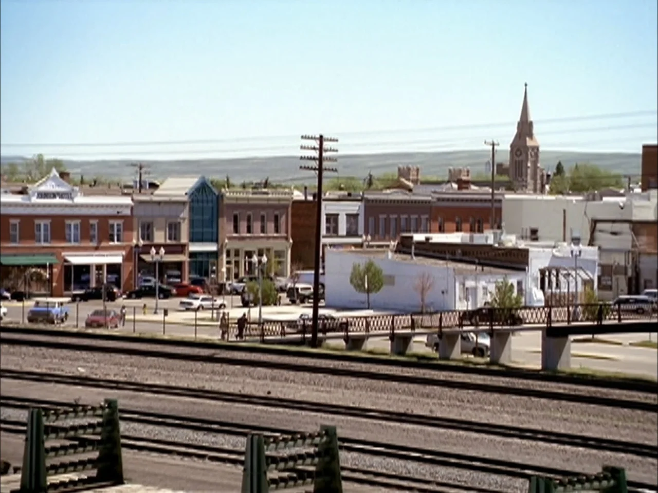 The Laramie Project (2020) movie still: establishing — A row of historic storefronts and a church steeple sit behind a train track. Parallel steel…
