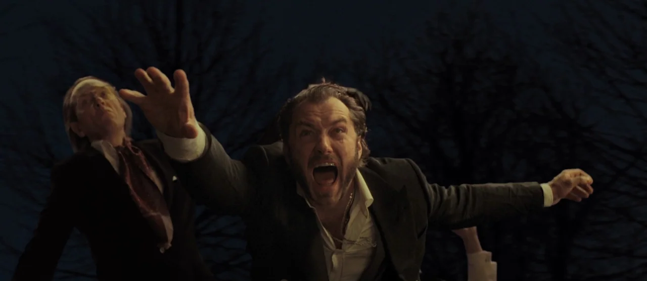 Dom Hemingway (2013) movie still: medium — A dark outdoor space with bare tree branches silhouetted against a night sky. Two men in formal business…