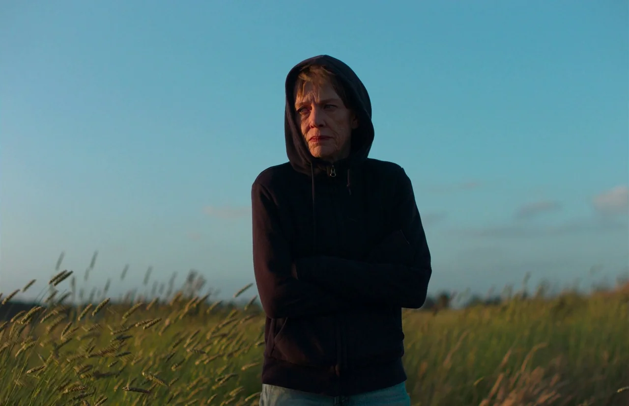 Nitram (2021) movie still: medium — A woman stands in a grassy field during the late afternoon. The sky is clear and blue.