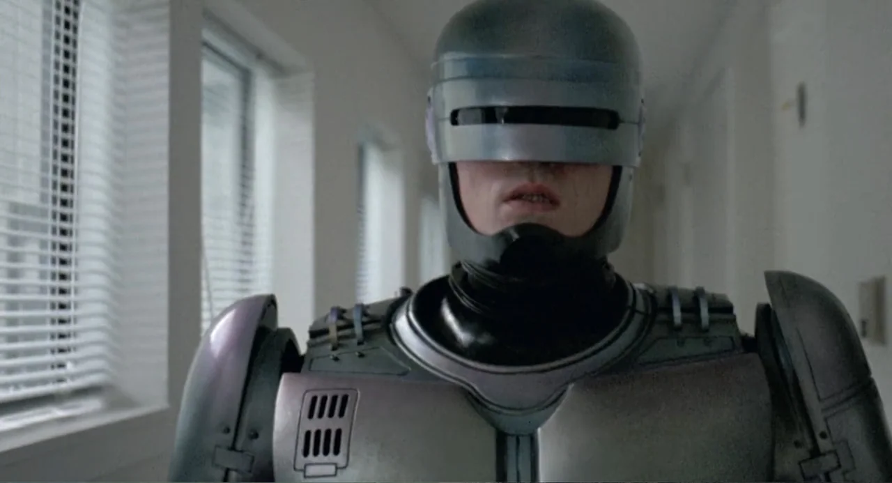 RoboCop (2014) movie still: medium — A sterile, white hospital hallway with light reflecting off windows featuring closed venetian blinds. A cyborg…