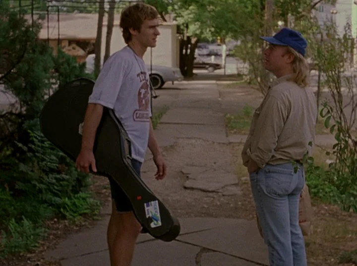 Slacker (1991) movie still: fullbody — A sidewalk runs through a residential area with lush green foliage and parked cars. Two young men stand…