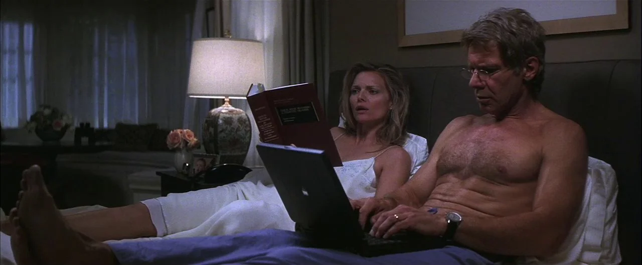 What Lies Beneath (2000) movie still: medium — A dimly lit bedroom features a large headboard and a bedside lamp. A couple sits in bed, the man…