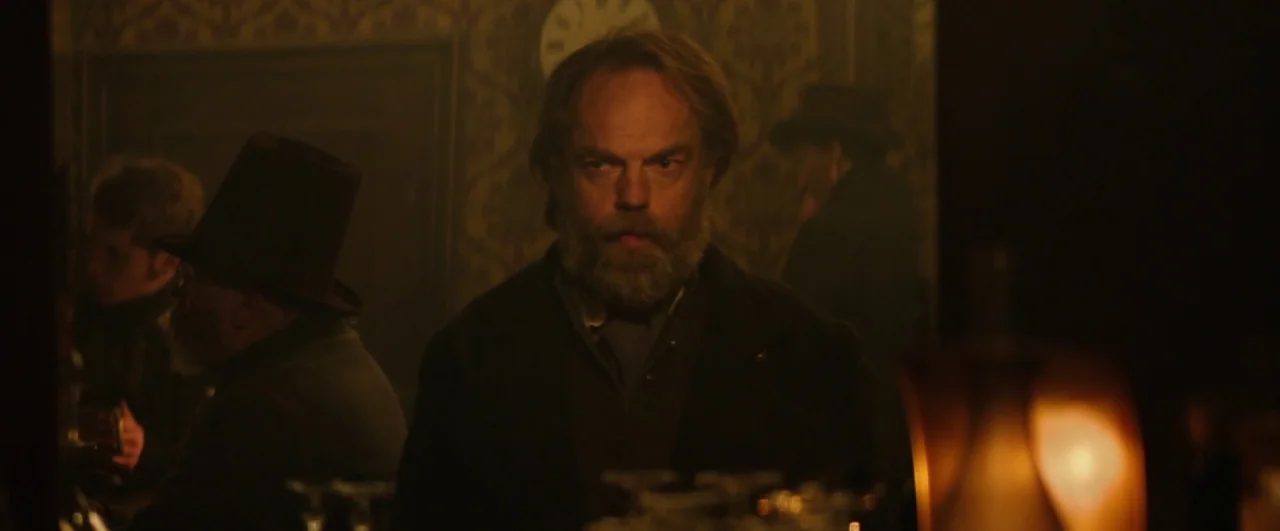 Black ’47 (2018) movie still: medium — A dimly lit tavern with patterned wallpaper and an analog clock on the wall. A man with a full beard stands…