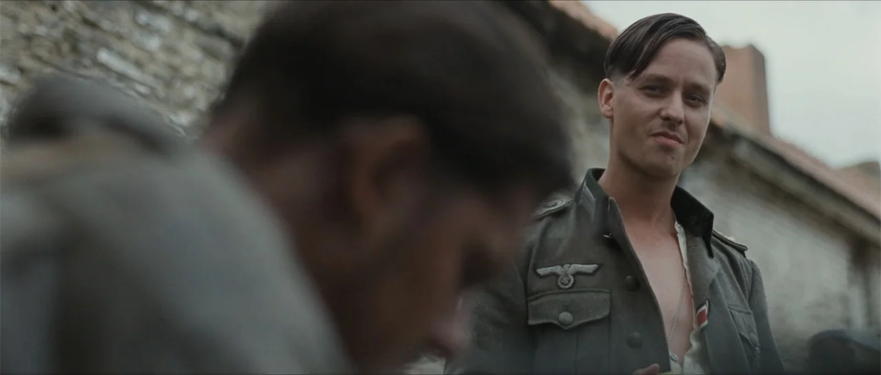 Suite Française (2015) movie still: medium — A man in a military jacket stands in front of a rustic stone building. A soldier in uniform watches a…