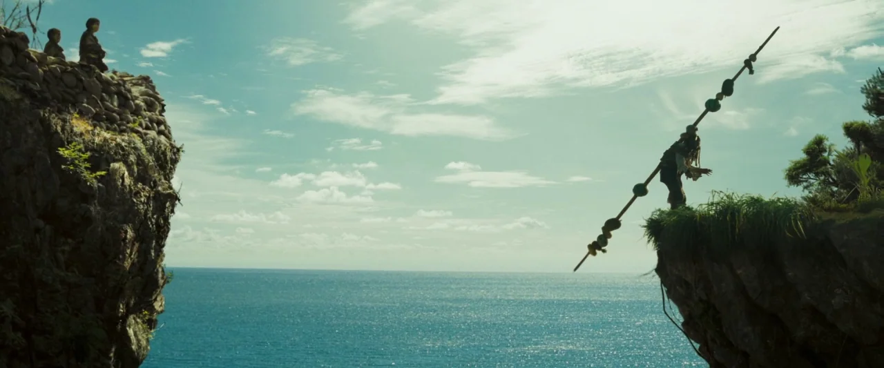 Pirates of the Caribbean: Dead Man’s Chest movie still: wide — Two rugged rock formations frame a vast expanse of bright blue ocean. A man stands on…