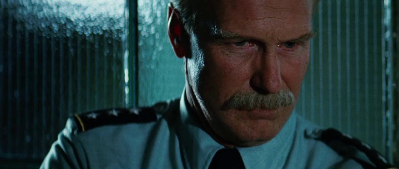 The Incredible Hulk (2008) movie still: closeup — An older military officer stands in a dimly lit office against a ribbed glass wall. The man peers…