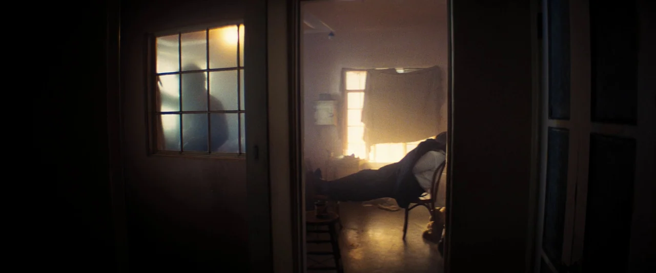 Babylon (2022) movie still: medium — A dimly lit room with a person slumped in a chair facing a window. Another figure stands in silhouette behind a…