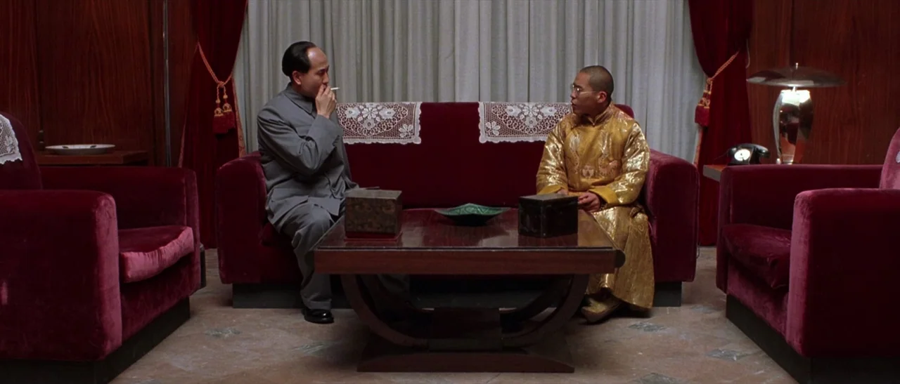 Kundun (1997) movie still: wide — A formal lounge area features deep red velvet chairs and matching couch against white curtains. Two men in…