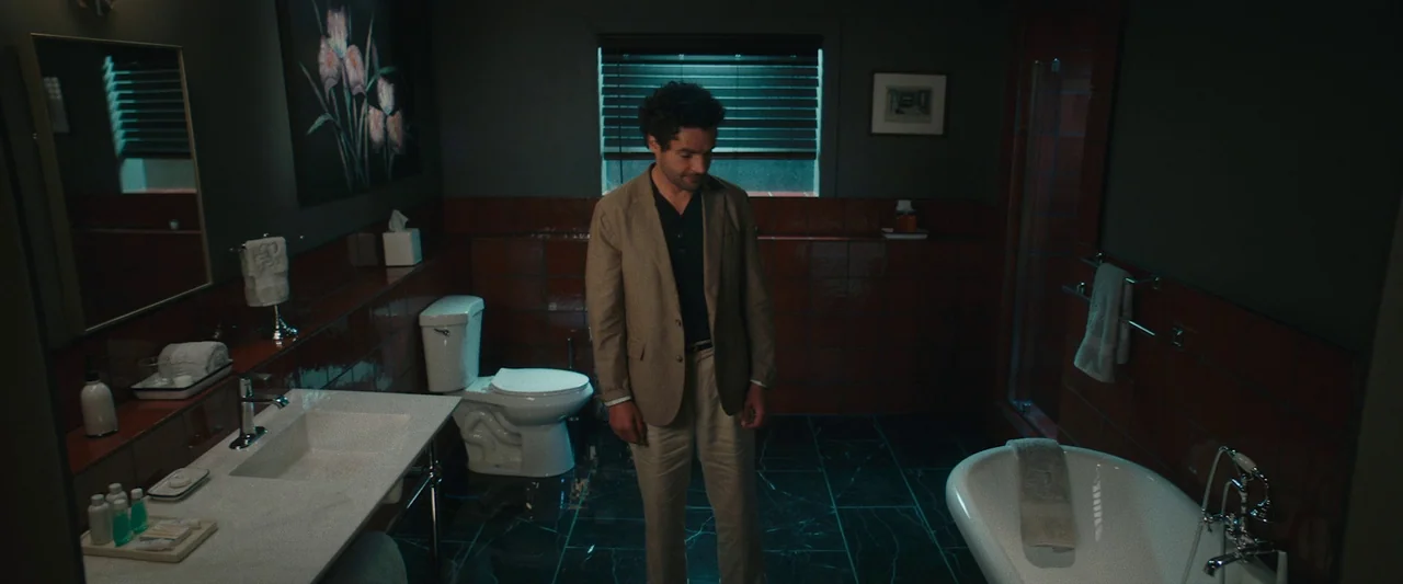Sanctuary (2023) movie still: fullbody — A modern bathroom features dark walls, glossy maroon tiles, and dark marble flooring. A man stands in the…