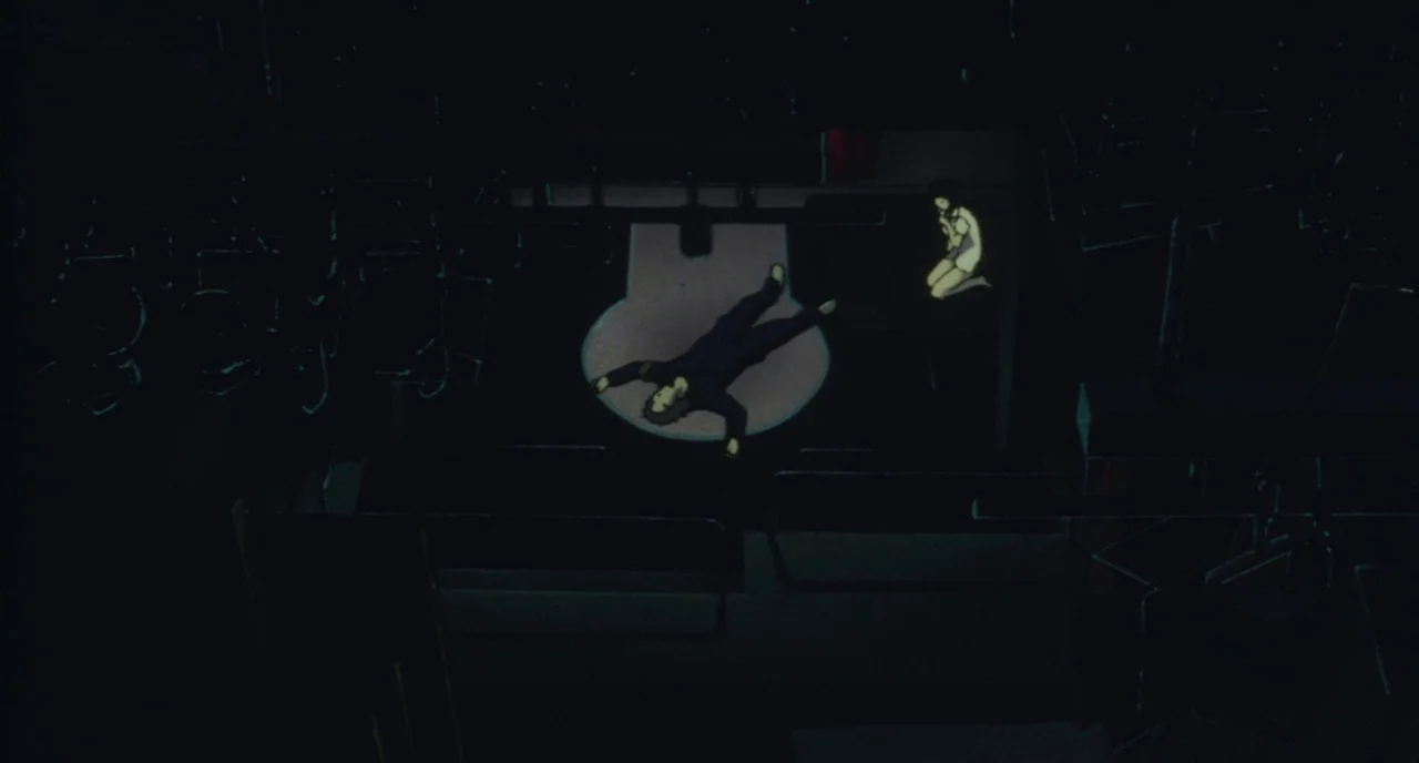 Perfect Blue (1997) movie still: wide — A dark, empty space resembling an auditorium with rows of seating visible in the darkness. A man lies on the…