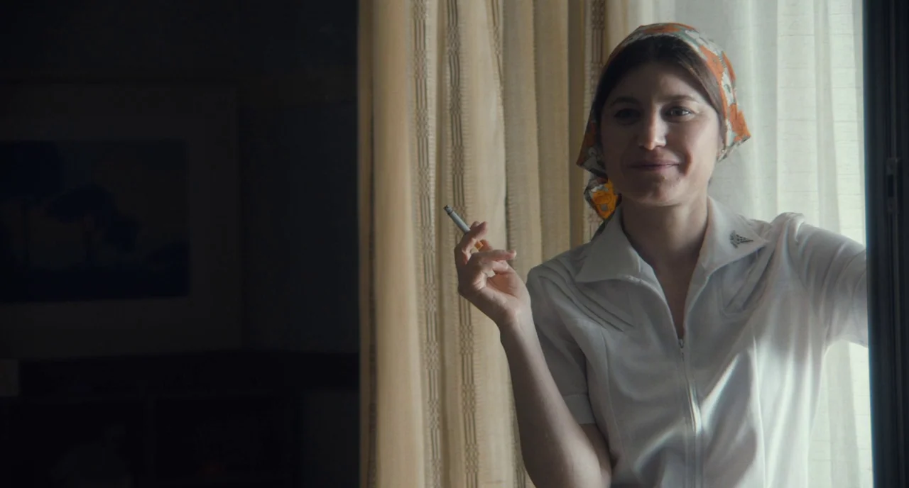 Christine (2016) movie still: medium — A woman stands indoors beside a window covered by beige curtains. She holds a cigarette while facing toward…