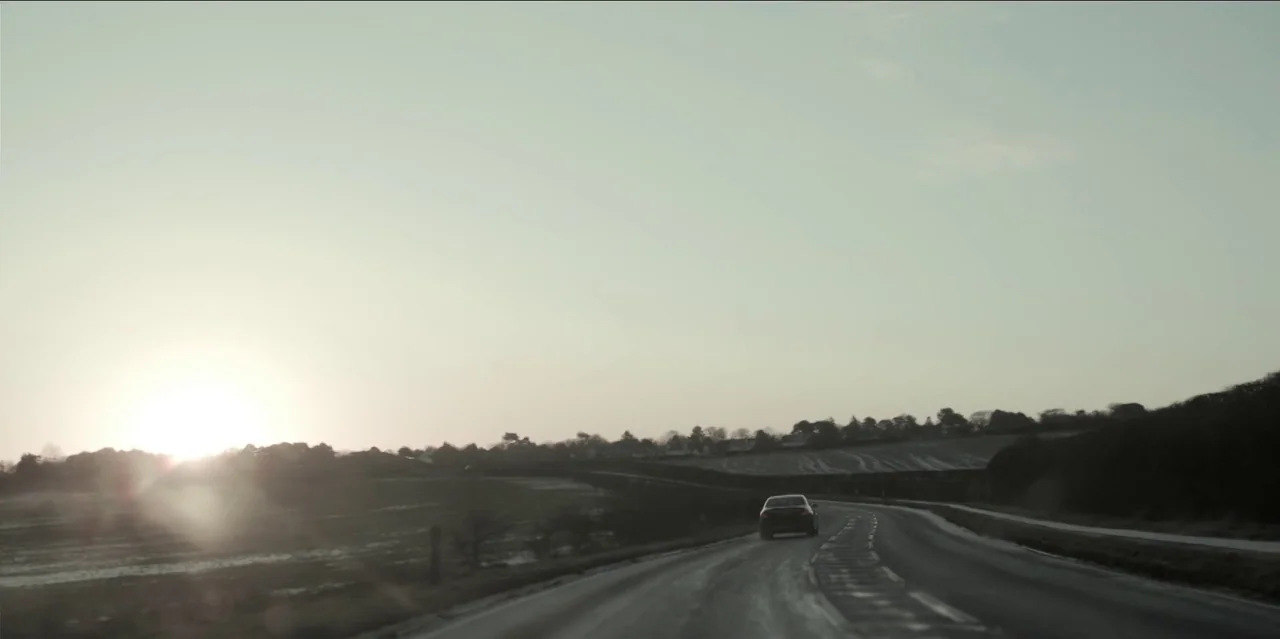 Help (2021) movie still: wide — A rural paved road winds through flat, open fields during a pale sunset. A single dark car travels away from the…