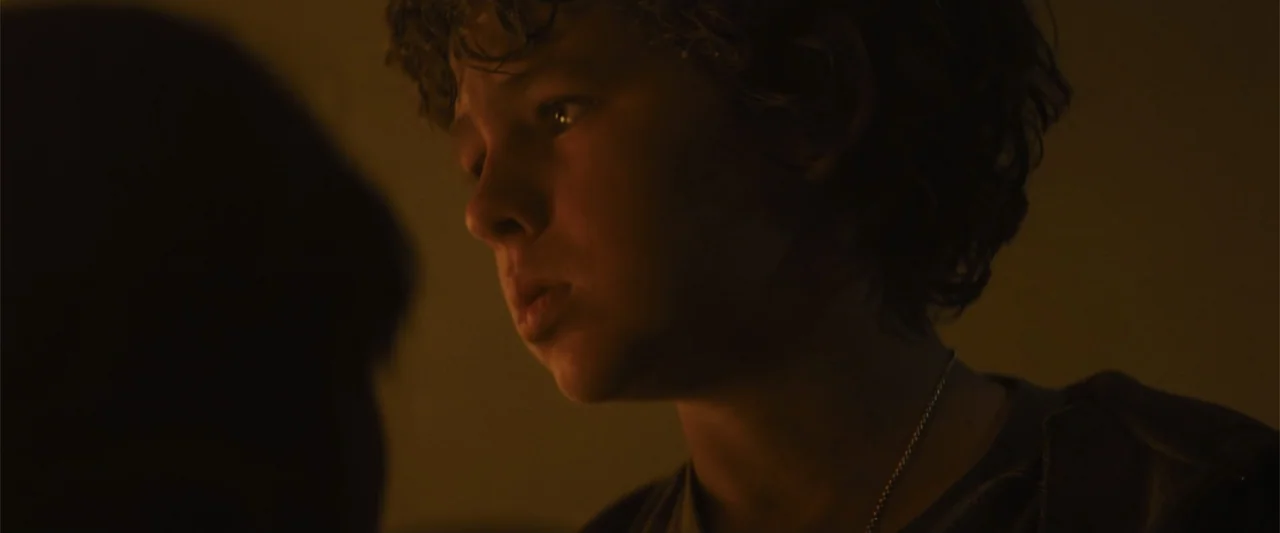 Those Who Wish Me Dead (2021) movie still: closeup — A dimly lit indoor space with low-key warm illumination. A young boy looks toward an unseen…