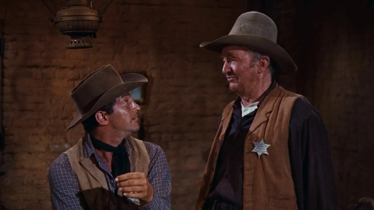 Rio Bravo (1959) movie still: medium — The interior of a rustic, dimly lit western space with brown brick walls. Two men in cowboy hats and suede…