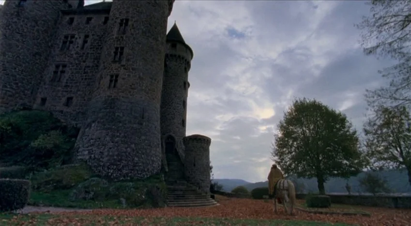 Bluebeard (2009) movie still: wide — A stone castle with high towers looms over a leaf-covered courtyard during autumn. A lone rider on a horse…