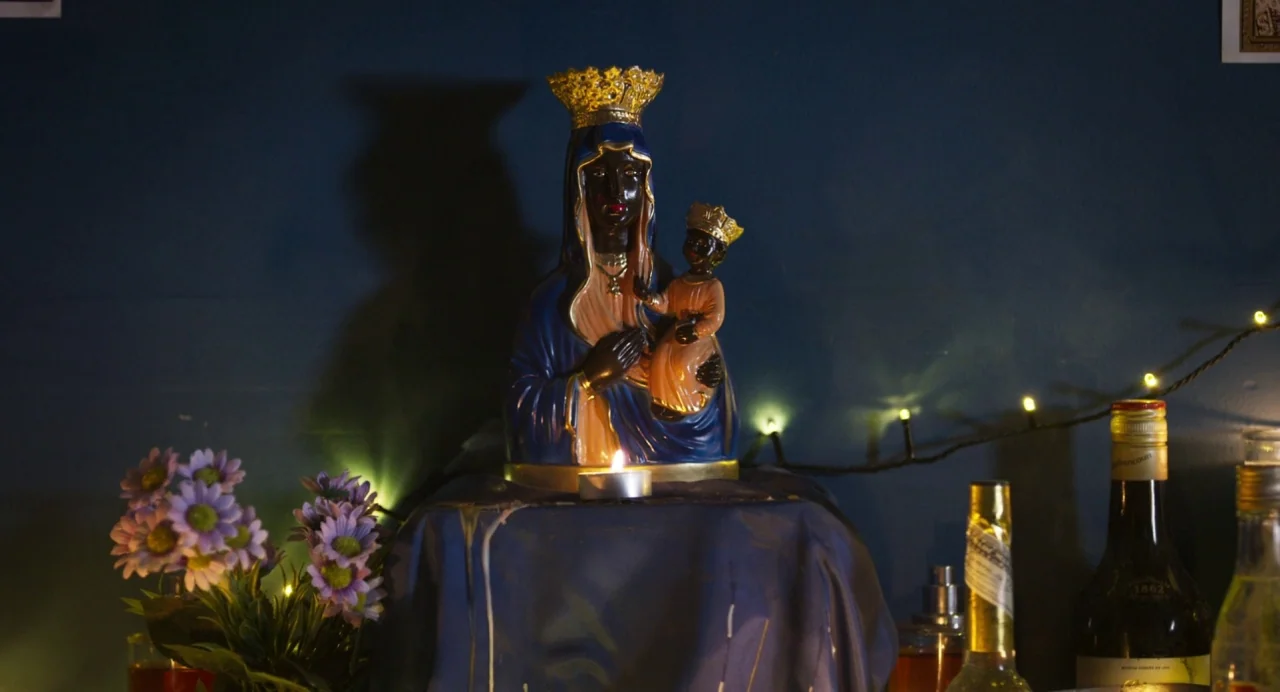 Zombi Child (2019) movie still: medium — A dark blue wall serves as the backdrop for a religious altar setup. A ceramic figurine of the Virgin Mary…