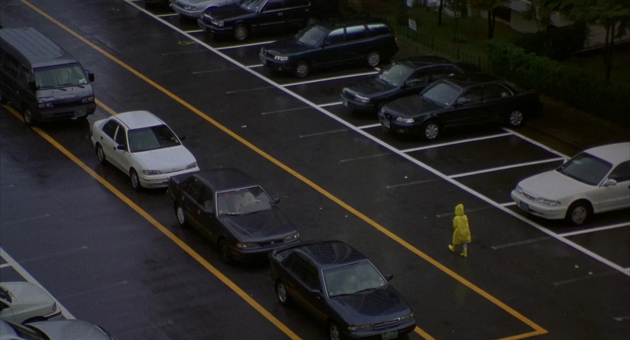 Barking Dogs Never Bite (2000) movie still: wide — An expansive asphalt parking lot is filled with numerous parked cars under overcast skies. A…