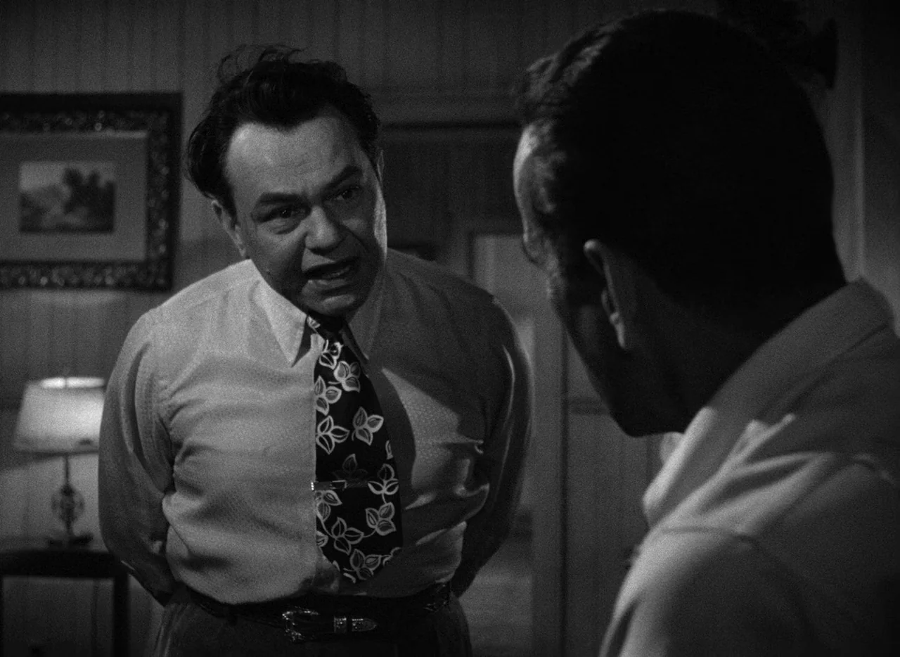 Key Largo (1948) movie still: medium — A dimly lit room features a man speaking to another person just out of frame. The man stands with hands…