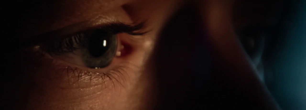 28 Years Later (2025) movie still: closeup — A extreme close-up of a human eye in deep shadow. A faint blue glow illuminates the side of the iris.