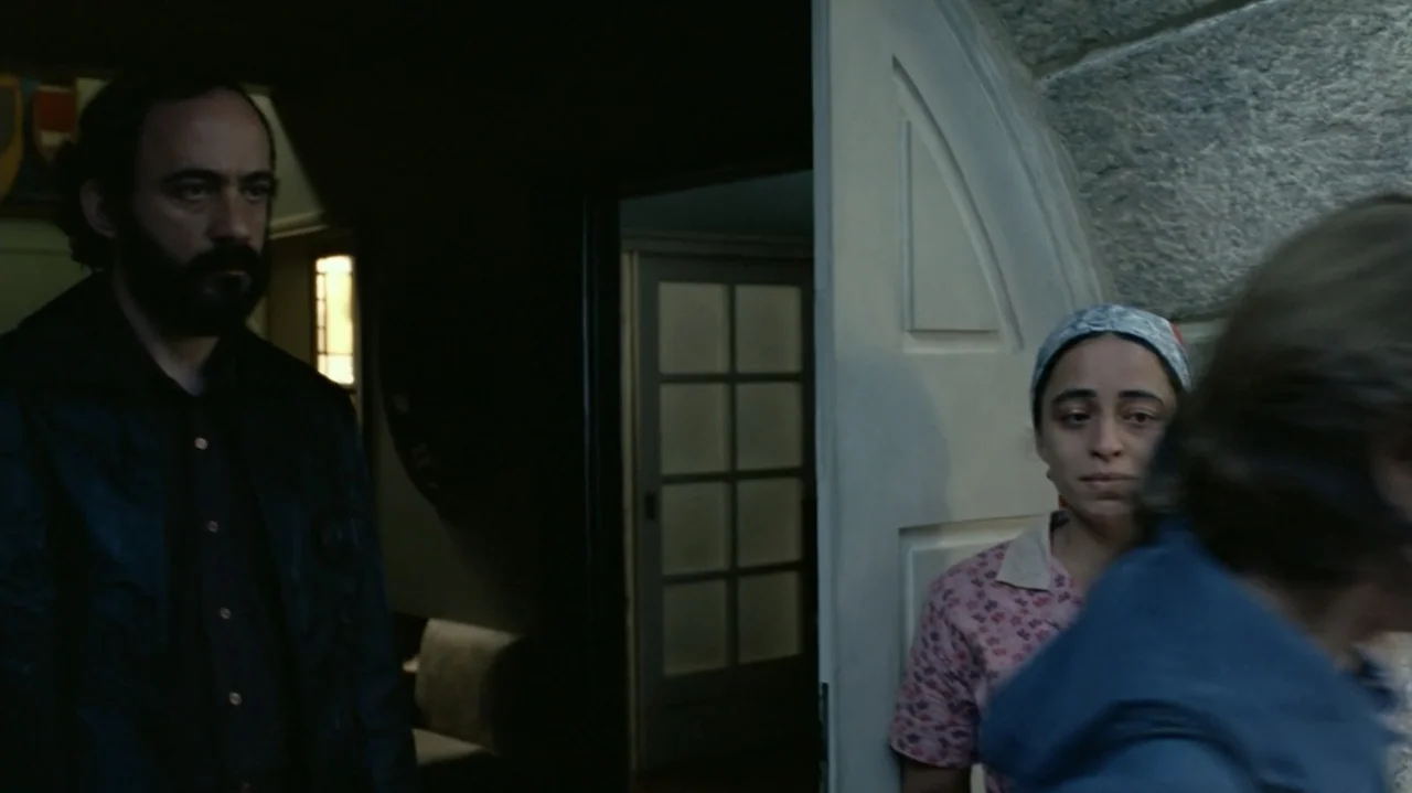 I'm Still Here (2024) movie still: medium — A man and a woman stand by an open door in an interior space. The woman wears a patterned headscarf and…