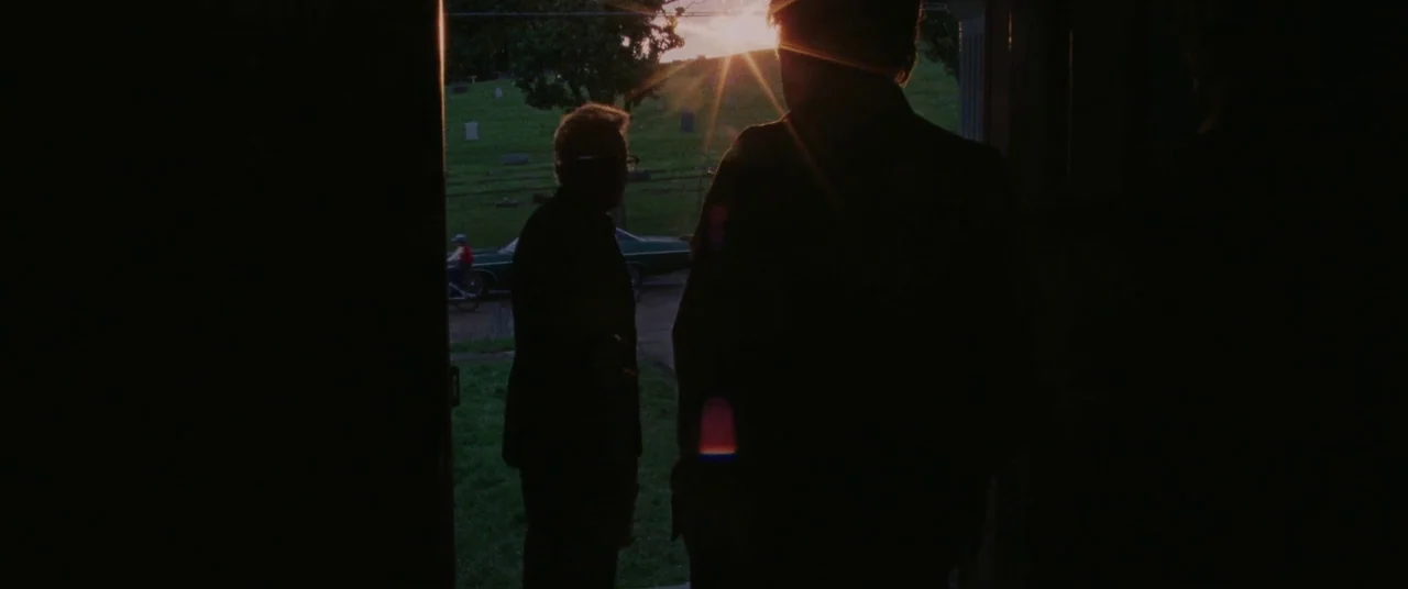 The Old Man & The Gun (2018) movie still: medium — A view from inside a dark doorway looking out toward a cemetery at sunset. Two men stand in the…
