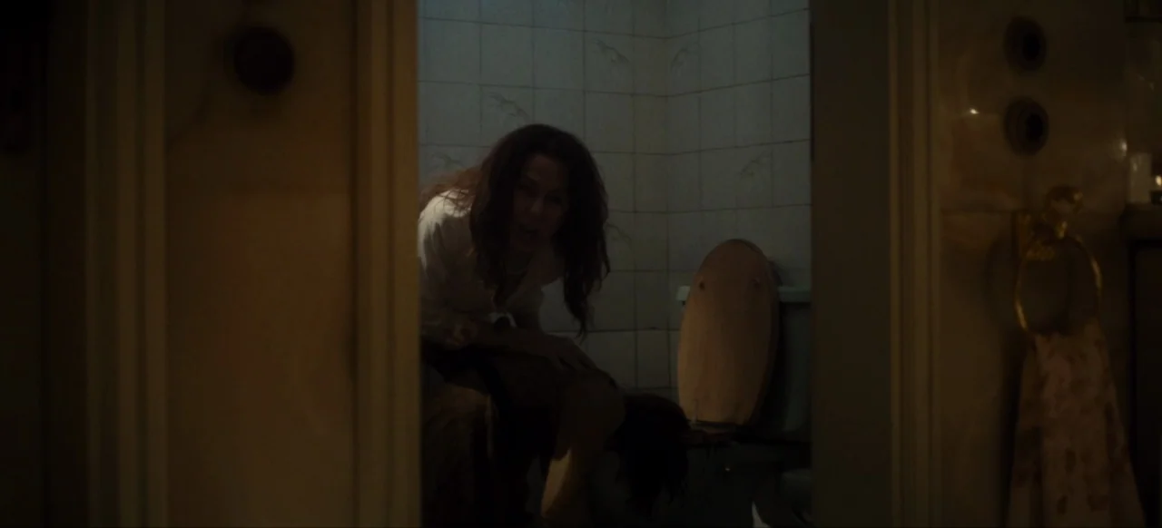 Housewife (2017) movie still: medium — A dimly lit room with light-colored wall tiles and a wooden toilet seat cover. A woman with long wavy brown…