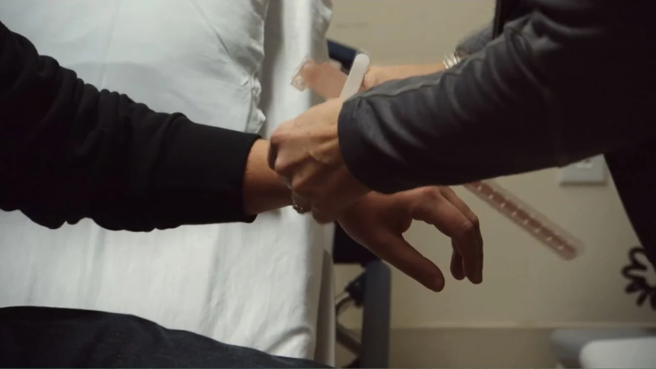 Suited (2016) movie still: closeup — A clinical medical examination room features white walls and a paper-covered exam table. One person applies a…