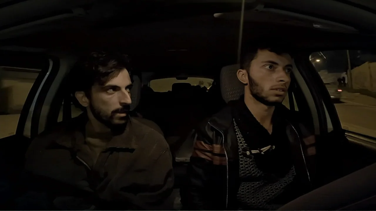 No Other Land (2024) movie still: medium — A dark car interior at night shows two men in the front seats. The driver and passenger look ahead while…
