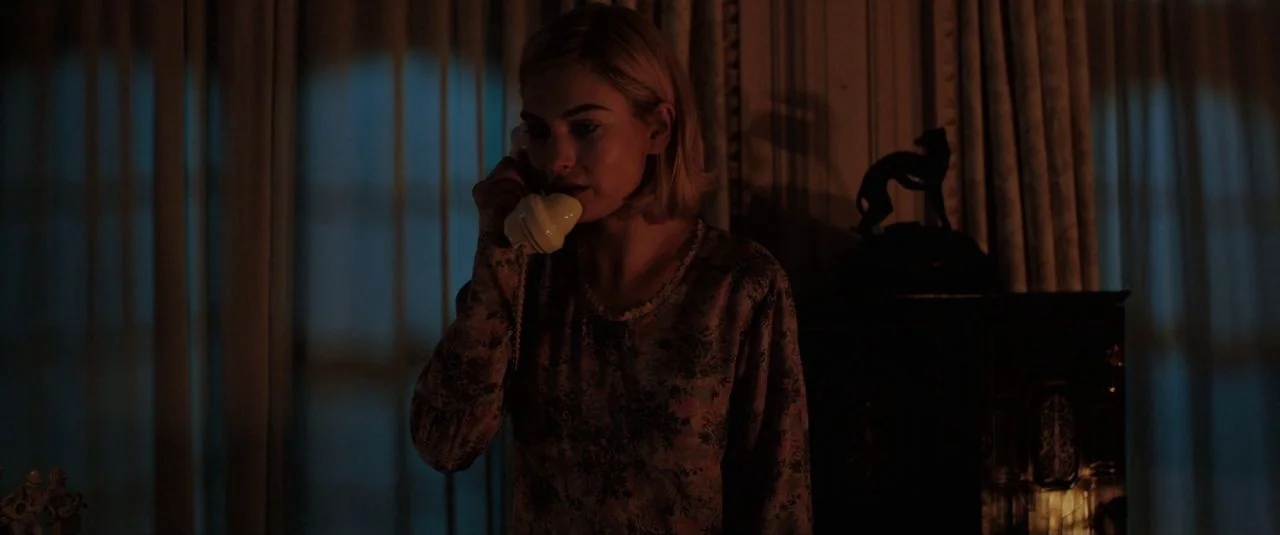 Rebecca movie still: medium — A woman stands in a dim room with drawn heavy curtains and dark wood furniture. She holds an off-white landline…