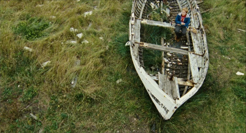 This Is England (2006) movie still: wide — An abandoned, weathered wooden boat sits amidst tall wild grasses in a meadow. A young boy sits inside…