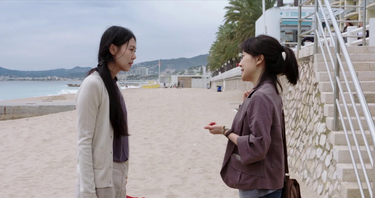 Claire’s Camera (2017) movie still: medium — A sandy beach area runs alongside a stone-walled embankment with stairs and palm trees. Two women stand…