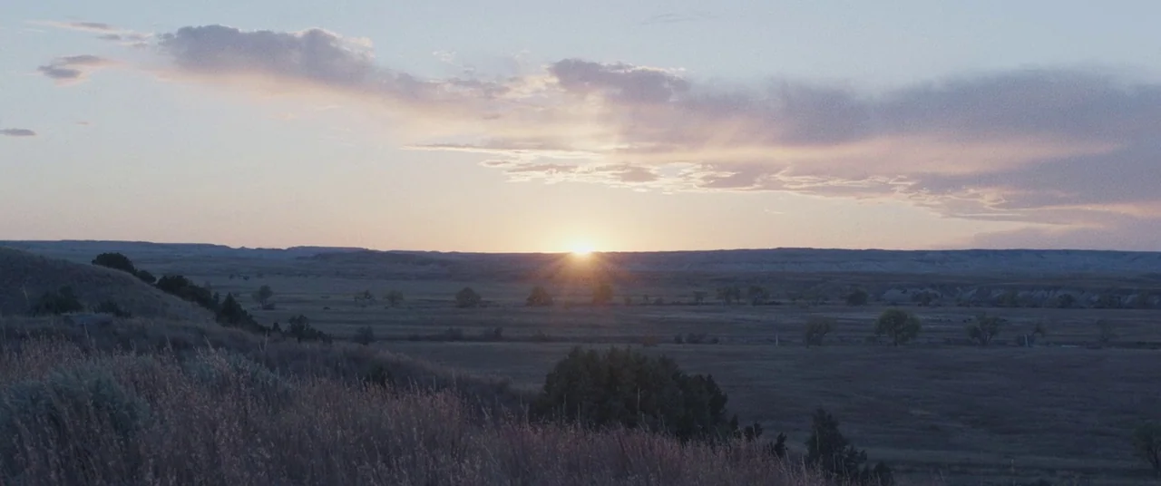 The Rider (2017) movie still: establishing — An expansive prairie landscape stretches toward a distant ridge under a soft sky. The sun emerges above…