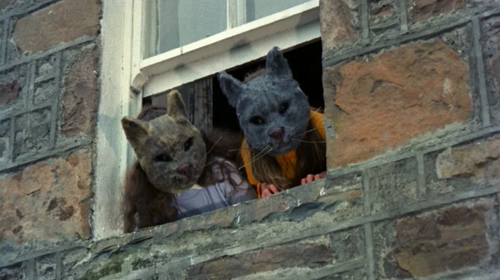 The Wicker Man movie still: medium — A stone building exterior features a rectangular window framed by rough-hewn masonry blocks. Two small figures…