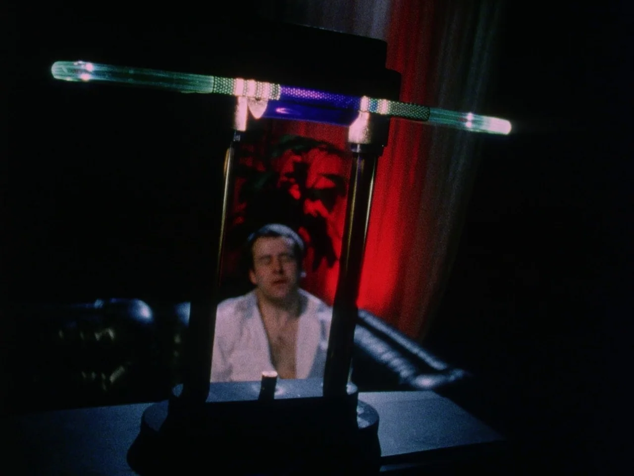 Subconscious Cruelty (2000) movie still: medium — A dimly lit lounge features a dark leather sofa against a red backdrop. A man in an unbuttoned…