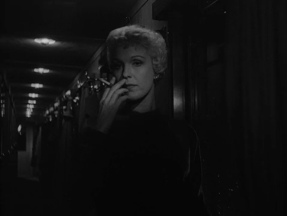 Dreams (1955) movie still: medium — A dark narrow train corridor features receding ceiling lights and shadowy walls. A blonde woman stands in the…