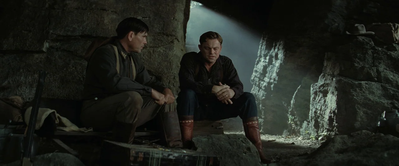 Killers of the Flower Moon (2023) movie still: medium — A dark rocky cave interior features large boulders and scattered camping gear items. Two men…