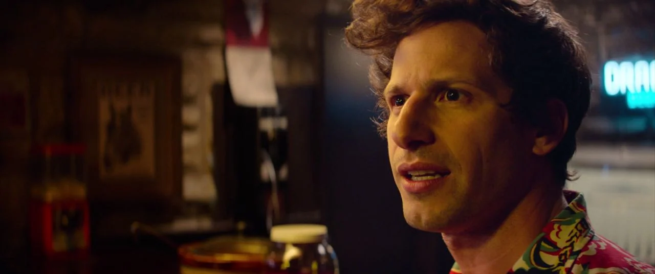 Palm Springs (2020) movie still: closeup — The interior of a bar with a dimly lit background featuring a blurred neon sign. A man with wavy hair…