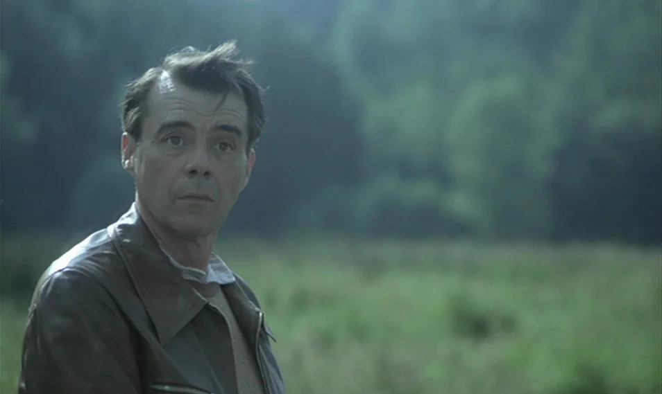 Despair (1978) movie still: medium — An open green meadow with blurred trees in the background under daylight. A middle-aged man in a leather jacket…