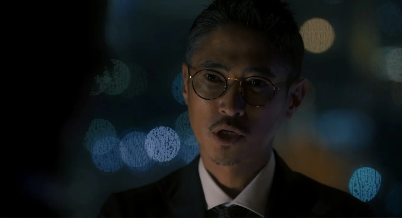 Knuckle Girl (2023) movie still: closeup — A man in a suit stands in a dark room at night. He speaks while out-of-focus city lights appear behind him.