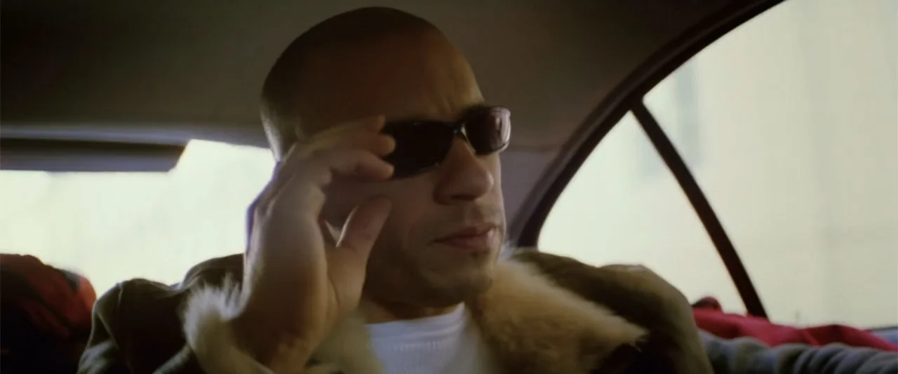xXx (2002) movie still: medium — A man sits in the backseat of a moving vehicle during the day. The subject adjusts his sunglasses while looking…