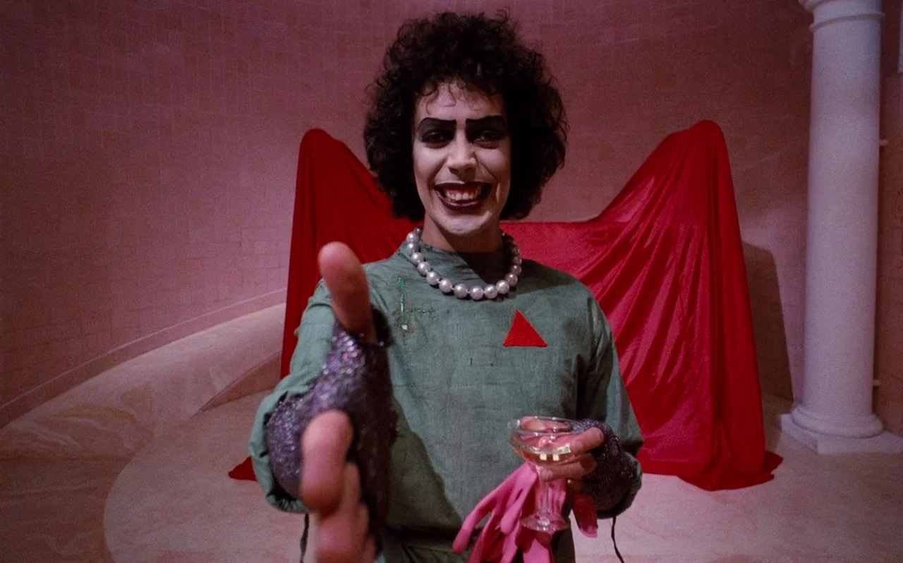The Rocky Horror Picture Show (1975) movie still: medium — A stone room with a tiled wall features a large red fabric draped behind. A person with…