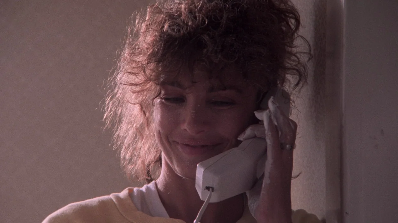 Fatal Attraction (1987) movie still: closeup — A woman stands indoors against a plain wall with dust on her skin. She is speaking on a telephone…